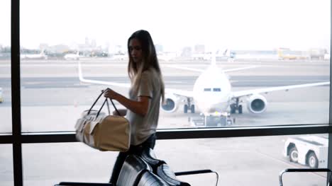 European-female-tourist-walks-up-to-airport-lounge-terminal-window,-puts-her-bag-down-and-looks-at-airplanes-passing