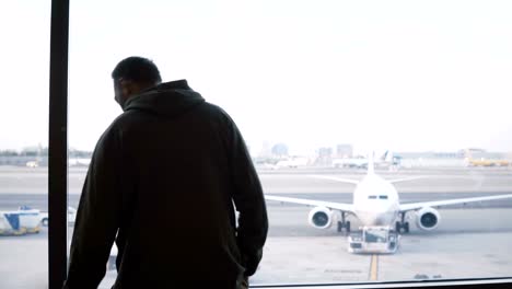 Male-traveler-with-big-bag-on-shoulder-walking-up-to-airport-lounge-terminal-window-to-enjoy-the-view-of-planes-and-cars