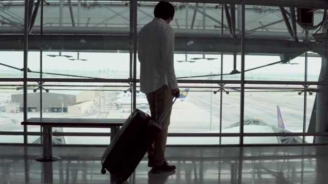 4-pasajeros-K.-macho-joven-con-smartphone-caminando-con-el-equipaje-de-la-maleta-y-sentado-sobre-un-banco-en-la-zona-de-salida-de-la-terminal-del-aeropuerto.-Empresario-asiático-en-viaje-de-negocios.-Conceptos-de-estilo-de-vida-moderno-viaje