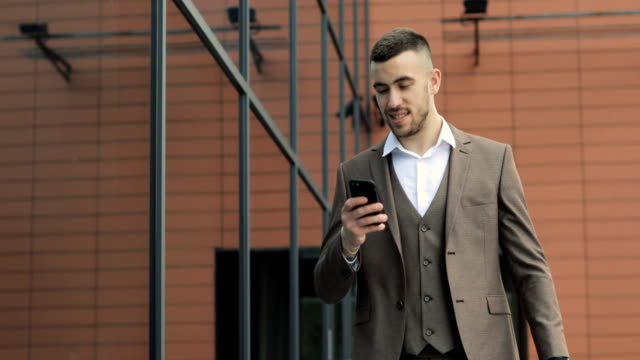 Mann-mit-Business-app-auf-Smartphones-zu-Fuß-in-die-Stadt.-Hübscher-junger-Geschäftsmann-Kommunikation-auf-Smartphone-lächelt-zuversichtlich