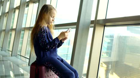 Entzückende-kleine-Mädchen-im-Flughafen-in-der-Nähe-von-großen-Fenster,-die-mit-ihrem-Handy-spielen