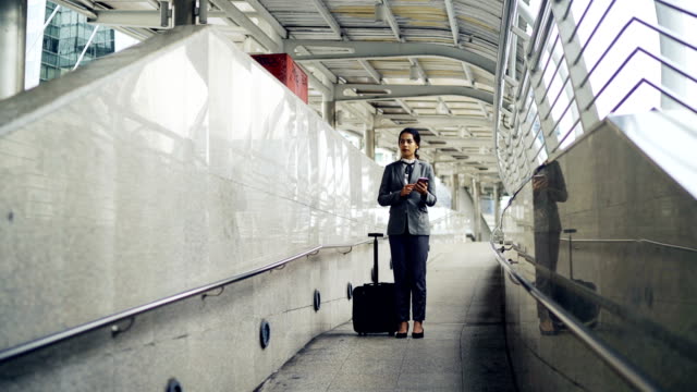 Asien-geschäftsfrau-im-Gespräch-des-Handys-während-des-Ziehens-einer-fahrbaren-Gepäcks-an-der-flughafenrampe-für-Geschäftsreise