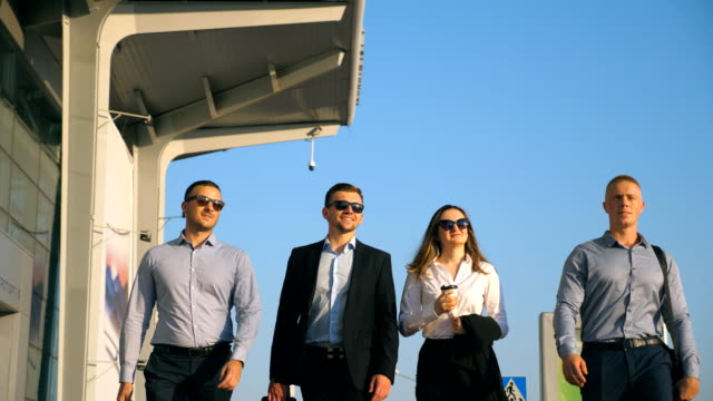 Group-of-young-happy-business-partners-walking-in-city-street-to-airport-terminal.-Colleagues-being-on-his-way-to-business-trip.-Portrait-of-smiling-business-people-stepping-and-talking.-Slow-motion-Close-up