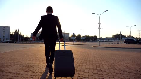 Unrecognizable-businessman-in-black-suit-is-late-for-the-flight-and-quickly-going-with-his-suitcase-to-airport.-Young-man-looking-at-his-watch-and-in-hurry-walking-with-luggage-to-terminal.-Rear-view