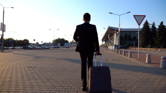 Dolly-shot-of-confident-young-man-in-a-formal-black-suit-walking-with-his-luggage-on-urban-street.-Successful-businessman-going-to-airport-terminal-and-pulling-suitcase-on-wheels-at-sunset.-Close-up