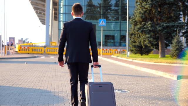 Rear-view-of-unrecognizable-young-businessman-walking-to-airport-terminal-and-pulling-suitcase-on-wheels-at-sunset.-Successful-male-business-person-going-with-his-luggage-on-city-street.-Close-up