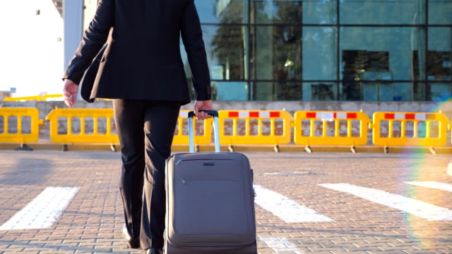 Dolly-shot-of-confident-young-man-in-a-formal-black-suit-walking-with-his-luggage-on-urban-street.-Successful-businessman-going-to-airport-terminal-and-pulling-suitcase-on-wheels-at-sunset.-Close-up