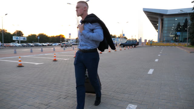 Happy-successful-businessman-walking-on-city-street-and-pulling-suitcase-on-wheels-after-flight-at-sunset.-Young-man-going-with-a-jacket-on-his-shoulder-against-the-background-of-airport.-Close-up