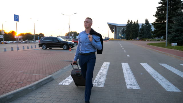Young-man-walking-on-urban-environment-with-a-jacket-on-his-shoulder-and-pulling-suitcase-on-wheels.-Successful-businessman-going-with-luggage-from-the-airport-on-city-street.-Close-up-Dolly-shot