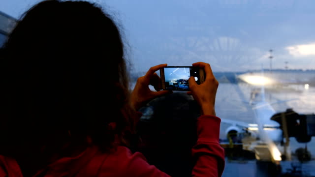 Mujer-en-una-sala-de-espera-del-aeropuerto-toma-una-imagen-de-un-plano-a-través-de-un-vidrio-a-un-primer-plano-de-smartphone
