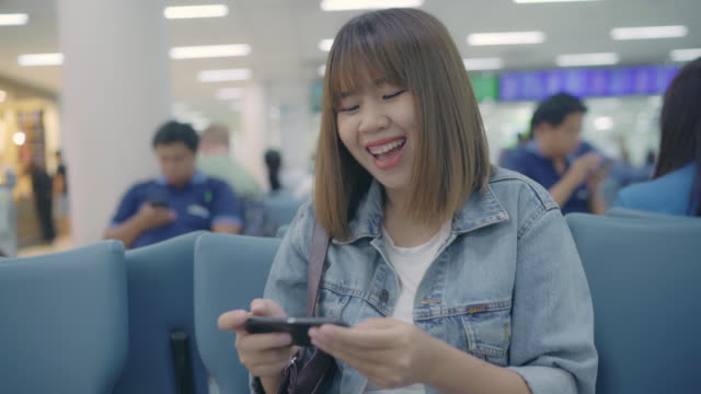 Feliz-mujer-asiática-utilizando-y-controlando-su-smartphone-sentado-en-silla-en-la-sala-terminal-mientras-espera-su-vuelo-en-la-puerta-de-salida-en-el-aeropuerto-internacional.-Mujer-feliz-en-concepto-de-aeropuerto.