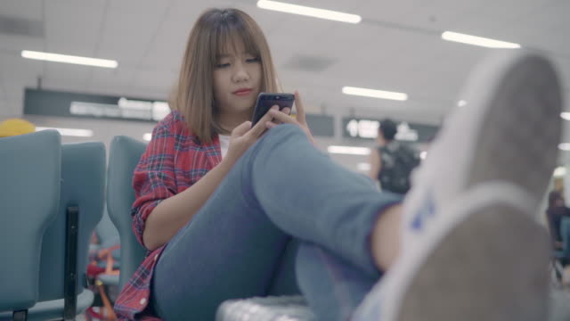 Happy-Asian-woman-using-and-checking-her-smartphone-while-sitting-on-chair-in-terminal-hall-while-waiting-her-flight-at-the-departure-gate-in-international-airport.-Women-happy-in-airport-concept.
