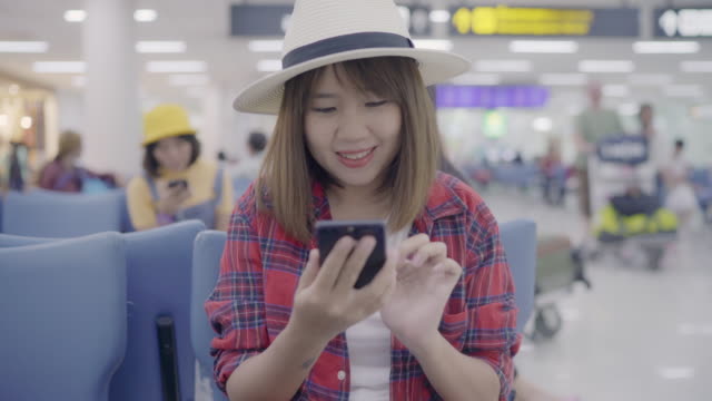 Gerne-Asiatin-mit-und-überprüfen-ihr-Smartphone-sitzend-auf-Stuhl-im-terminal-Halle-während-des-Wartens-ihr-Fluges-am-Abfluggate-im-internationalen-Flughafen.-Frauen-glücklich-im-Flughafenkonzept.