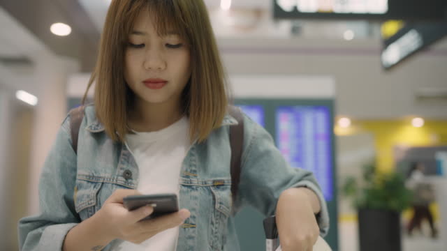 Feliz-mujer-asiática-utilizando-y-controlando-su-smartphone-en-el-hall-de-la-terminal-mientras-espera-su-vuelo-en-la-puerta-de-salida-en-el-aeropuerto-internacional.-Mujer-feliz-en-concepto-de-aeropuerto.