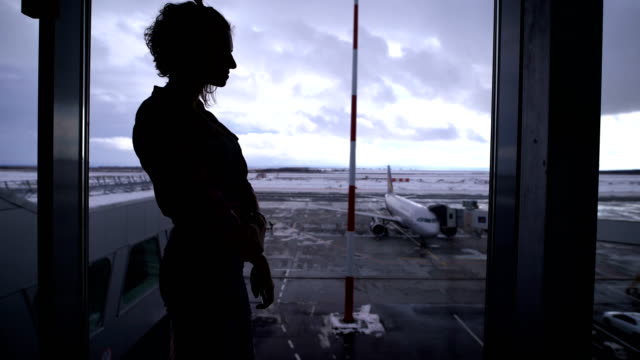 Girl-standing-at-the-airport-near-a-large-window-looking-out-on-the-planes-that-are-on-the-runway.-Silhouette-of-a-young-woman-against-the-gray-sky-and-standing-on-the-territory-aircraft