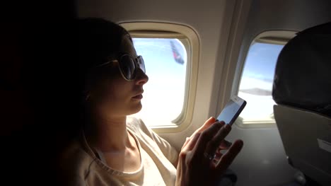 Tourist-woman-sitting-near-airplane-window-at-sunset-and-using-mobile-phone-during-flight