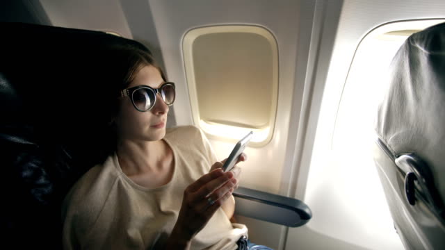 Tourist-Frau-sitzt-in-der-Nähe-von-Flugzeugfenster-bei-Sonnenuntergang-und-mit-Handy-während-des-Fluges