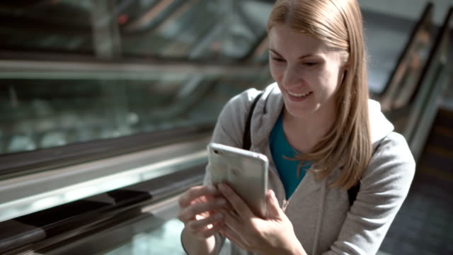 Mujer-alegre-con-escaleras-mecánicas-en-la-terminal-del-aeropuerto.-Hablar-con-amigo-en-smartphone-vía-Skype