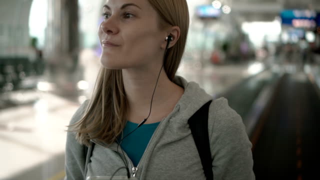 Feliz-sonriente-mujer-con-travolator-en-terminal-del-aeropuerto.-Con-su-smartphone,-escuchando-música