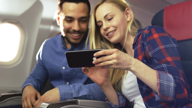 On-a-Board-of-Commercial-Airplane-Beautiful-Young-Blonde-with-Handsome-Hispanic-Male-Share-Videos-on-Smartphone-and-Smile.-Sun-Shines-Through-Aeroplane-Window.
