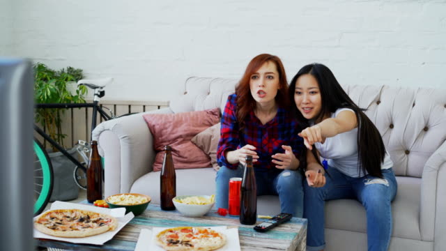Dos-amigas-multiétnicas-viendo-deportes-partido-en-TV-juntos-en-casa-bebiendo-cerveza-y-comiendo-snacks