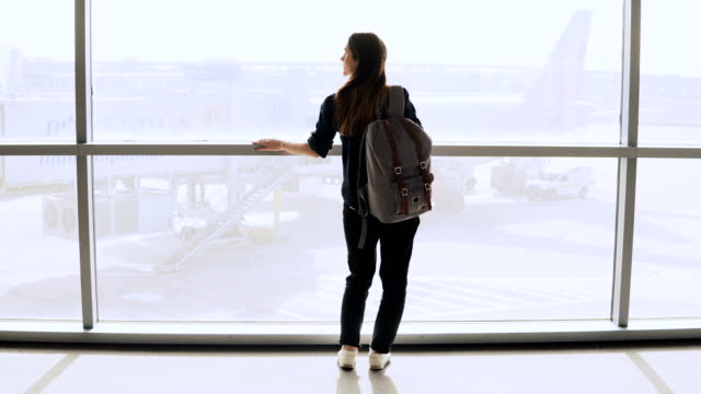 Young-woman-with-backpack-near-terminal-window.-Caucasian-female-tourist-using-smartphone-in-airport-lounge.-Travel.-4K