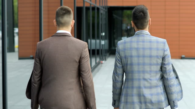 Two-businessmen-chat-together-as-they-walk-along-through-a-busy-modern-office-building.-Rear-view