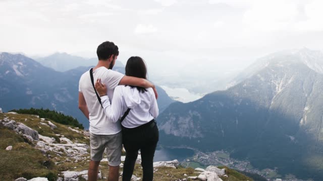 Joven-pareja-abrazándose-en-la-cima-de-la-montaña,-disfrutando-de-impresionantes-vistas-sobre-el-lago-de-las-montañas,-vista-posterior