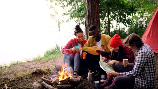 Cinemagraph-loop---smiling-guys-and-girls-are-looking-at-maps-resting-around-fire-in-woods-and-holding-drinks.-Flame-and-paper-is-moving,-beautiful-nature-is-visible.