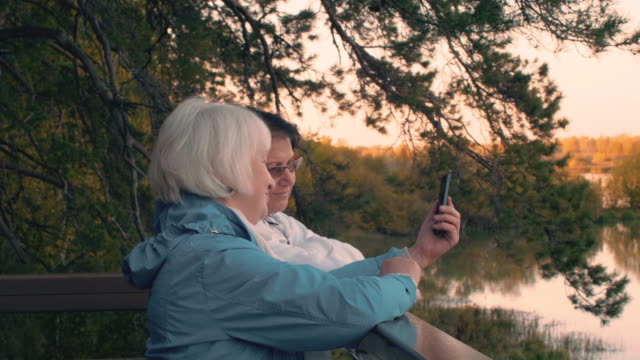Zwei-ältere-Frau-Herbst-Spaziergang-am-Baum-und-Seenlandschaft-Smartphone-betrachten