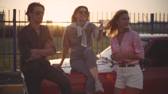friends-sit-on-the-hood-of-a-car-talk-and-laugh-at-sunset.-Car-trip