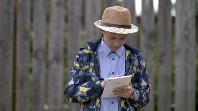 farmer-boy-in-a-hat-uses-a-tablet,-stands-against-the-fence