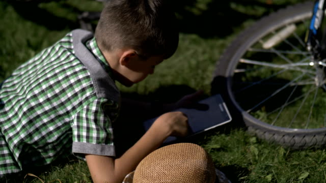 niño-se-encuentra-en-la-hierba-con-una-tableta-después-de-un-paseo-en-bicicleta,-al-aire-libre