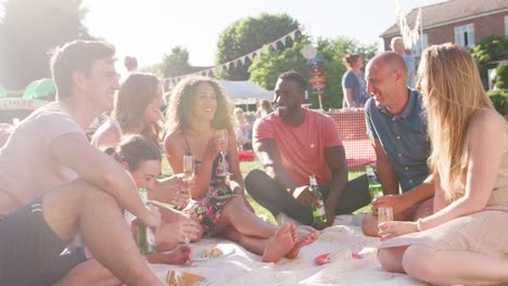 Group-of-friends-eating-and-drinking-as-they-sit-on-rug-at-summer-garden-fete---shot-in-slow-motion