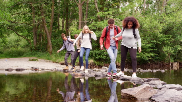 Five-young-adult-friends-hold-hands-and-help-each-other-while-crossing-a-stream-and-standing-on-stones-during-a-hike