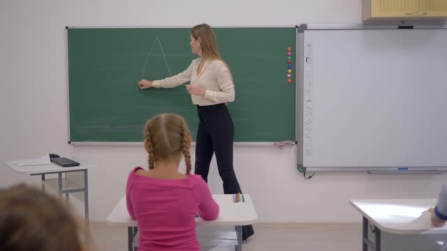 Junior-School,-mujer-del-profesor-dibuja-figuras-geométricas-a-bordo-durante-la-lección-para-los-escolares-sentados-en-pupitres-en-el-aula