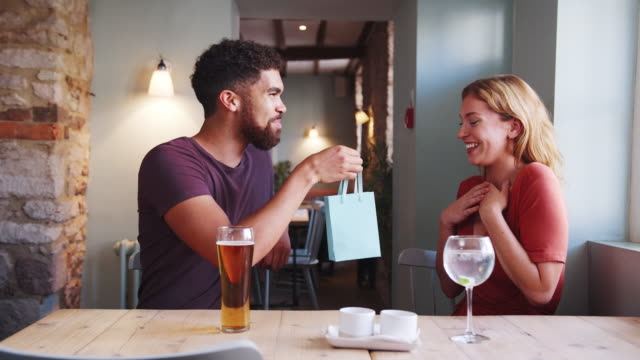 Happy-young-adult-woman-is-surprised-by-a-gift-from-her-boyfriend-while-they-sit-at-table-with-drinks-in-a-pub,-close-up