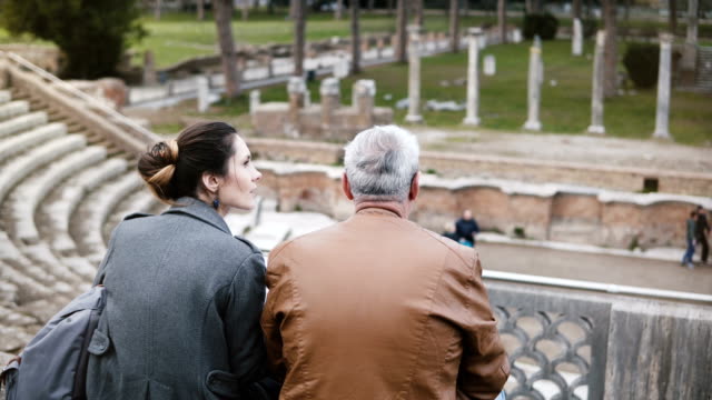 Rückansicht-des-Europäischen-Mädchen-und-ältere-Mann-sitzen-und-reden-an-alten-Ruinen-der-antiken-Ostia-Amphitheater-Italiens.