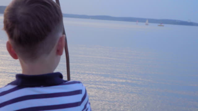 Caucasian-boy-in-a-striped-vest-on-the-waterfront.-Joyful-in-the-background-yachts,-sunset.
