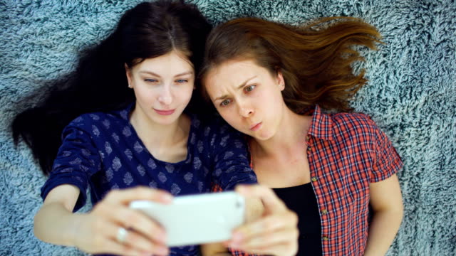 Top-view-of-two-pretty-girls-in-pajamas-making-selfie-portrait-on-bed-in-bedroom-at-home