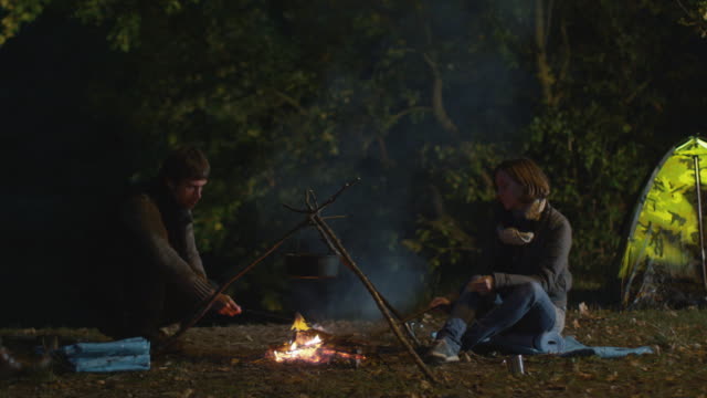 A-man-and-a-woman-sit-next-to-a-campfire-at-night-and-prepare-food-on-a-stick-while-another-male-throws-wood-in-the-fire.