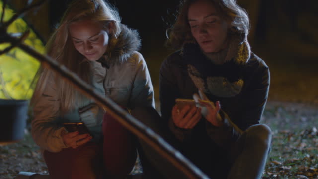 Zwei-glückliche-Mädchen-sitzen-nachts-neben-einem-Lagerfeuer-und-nutzen-ihre-Smartphones.