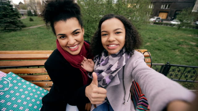 Zwei-niedliche-afroamerikanische-Frau-Selfie-auf-Smartphone-mit-Einkaufstüten-und-lächelnd.-Freundinnen-auf-Straße-Bank-sitzen-viel-Spaß-nach-dem-Besuch-der-Mall-Vertrieb