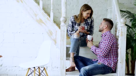 Young-happy-and-loving-couple-drink-tea-and-talking-while-sitting-on-stairs-in-living-room-at-home