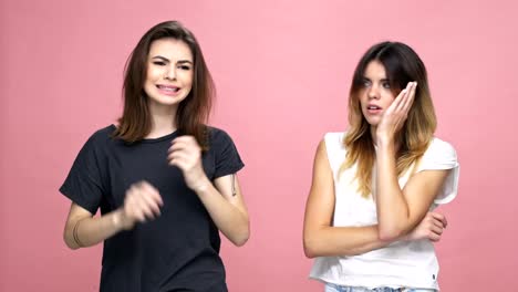 Two-upset-frustrated-girls-standing-together-and-gesturing-isolated-over-pink-background