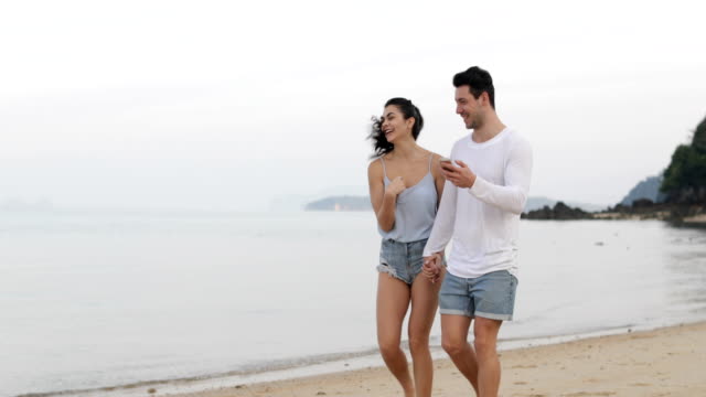 Paar-am-Strand-hält-Smart-Handys-sprechen,-junger-Mann-und-Frau-Touristen-Hand-in-Hand-gehen