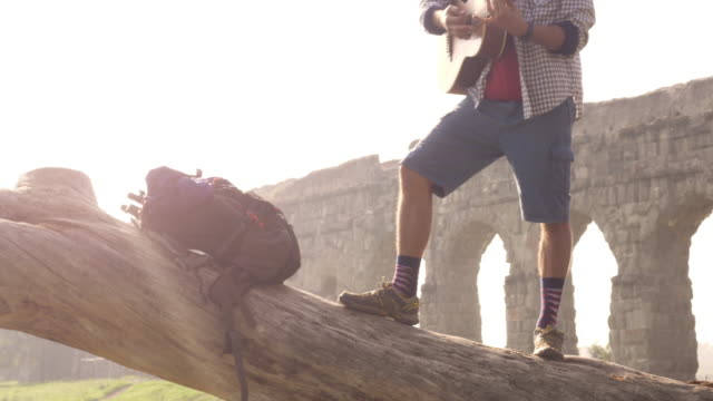 Young-man-adventurer-traveler-standing-on-top-of-a-log-trunk-playing-guitar-singing-in-front-of-ancient-roman-aqueduct-ruins-in-parco-degli-acquedotti-park-in-rome-at-sunrise-slow-motion
