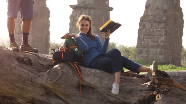 Happy-young-woman-backpacker-tourists-sitting-lying-on-a-log-trunk-reading-book-in-front-of-ancient-roman-aqueduct-ruins-in-parco-degli-acquedotti-park-in-rome-at-sunrise-romantic-couple-slow-motion