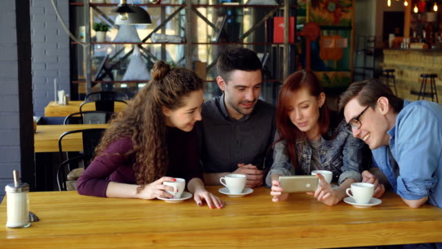 Good-looking-friends-are-watching-interesting-video-and-discussing-it-while-drinking-tea-at-table-in-cozy-cafe.-Modern-lifestyle-and-happy-people-concept.