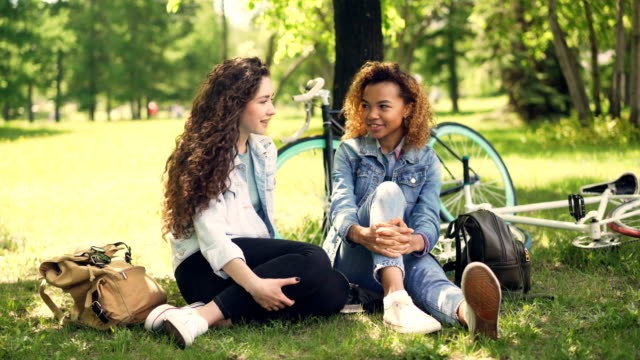 Cheerful-students-African-American-and-Caucasian-are-talking-and-laughing-sitting-in-park-on-lawn-after-riding-bikes.-Nature,-conversation-and-friendship-concept.